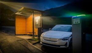 A white electric vehicle beside a public charging station at blue hour, with a wooden shelter and misty hills behind it.