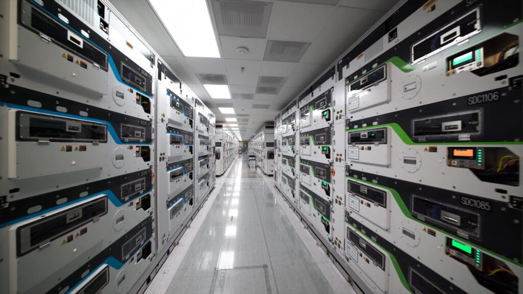 Aisle between tall rows of white and dark modular semiconductor test equipment cabinets with blue and green trim in a cleanroom facility.