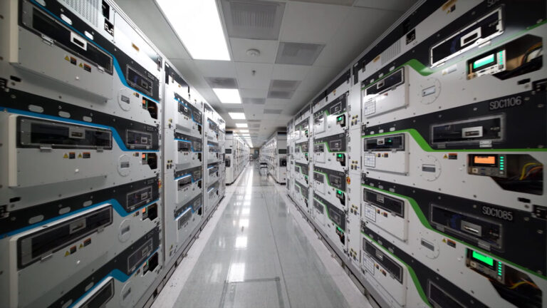 Aisle between tall rows of white and dark modular semiconductor test equipment cabinets with blue and green trim in a cleanroom facility.