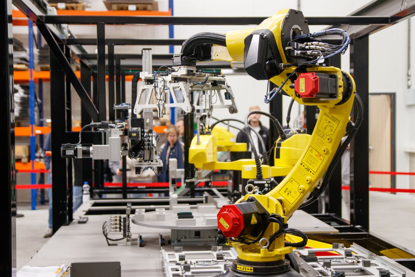 Yellow industrial robot arm with a gripper and connected cabling in an open-frame work cell, with other robots and people in the background.